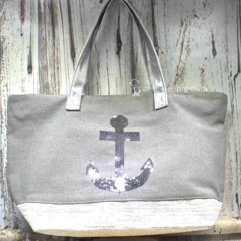 Graue Shoppertasche mit silbernem Pailletten-Anker, weißem Boden mit Silberstreifen und grauen Kunstleder-Griffen vor rustikalem Holzhintergrund.